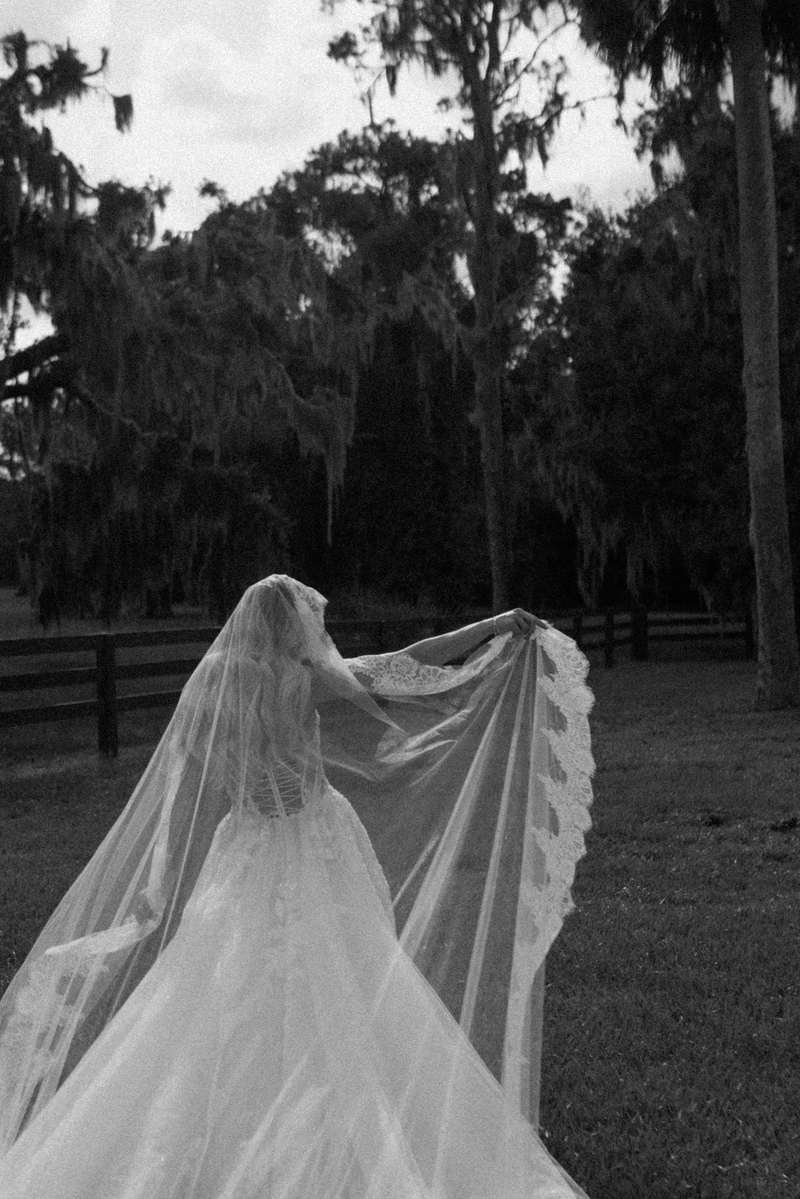 Black and white portrait of a bride holding out her long veil beneath moss-covered trees